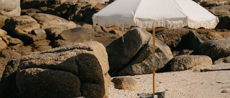 En strandscene med en hvid parasol, en strandstol, et håndklæde, en taske og en kurv i sandet.