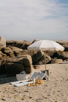 A beach setting with a white umbrella, a beach chair, a towel, a bag, and a basket on the sand.