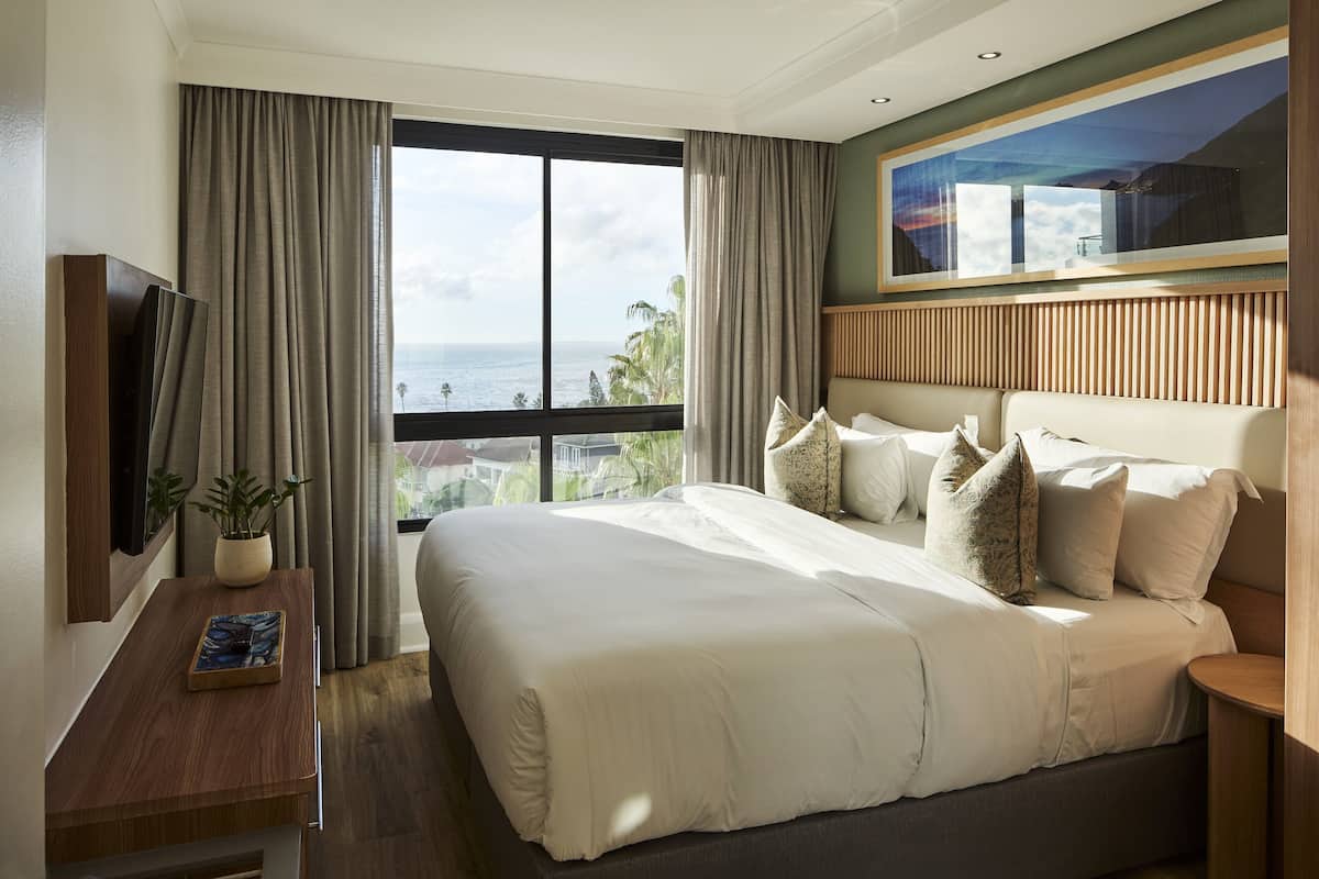 Ein Hotelzimmer mit einem großen Bett, einem an der Wand befestigten Fernseher, einem Fenster mit Vorhängen und Meerblick.
