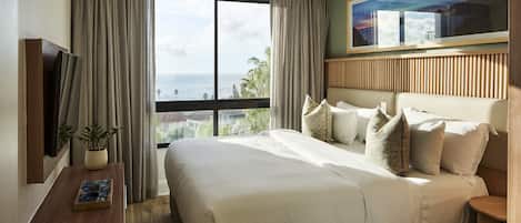 A hotel room with a large bed, a TV on the wall, a window with curtains, and a view of the ocean.