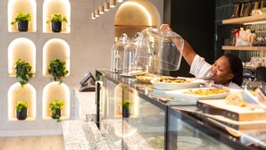 A buffet area with food displayed under glass covers, a person serving food, and potted plants in the background. - President Hotel (Cape Town)