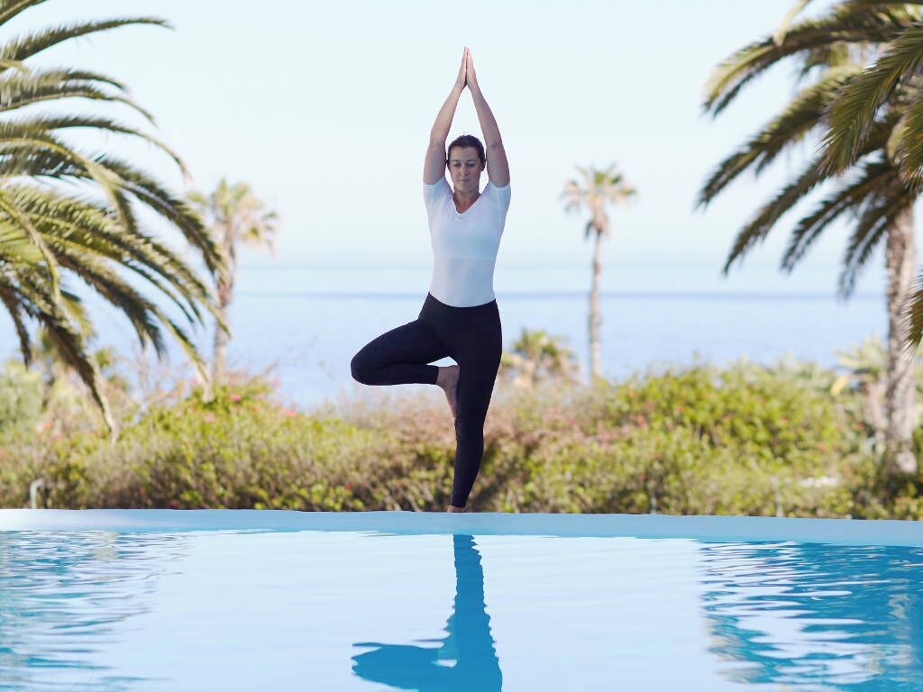A person performing a yoga pose by a pool with palm trees and a clear sky in the background.