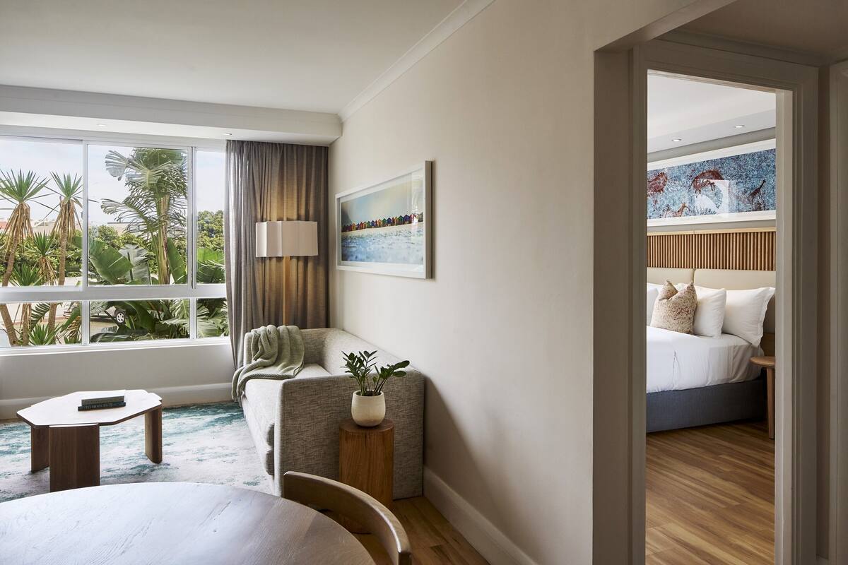 A hotel room with a large window, a sofa, a small table, and a bed with a wooden headboard.