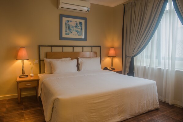 A neatly made bed with white linens, two bedside lamps, a framed artwork above the bed, and a window with sheer curtains.