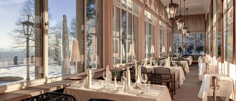 A restaurant with tables set for dining, large windows, and hanging light fixtures.