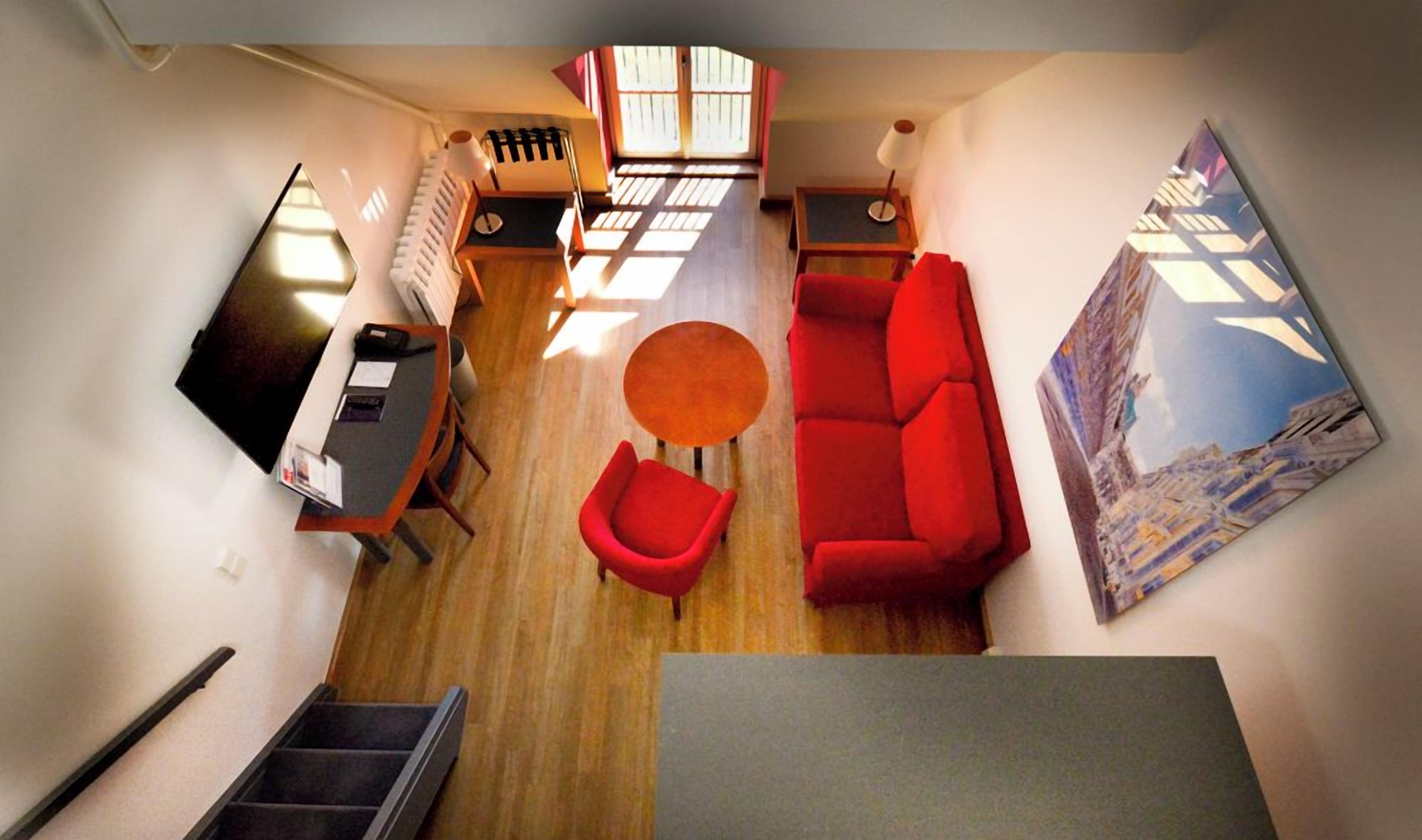 A compact living space with a red sofa, a small round table, a red armchair, a desk with a computer, and a radiator.