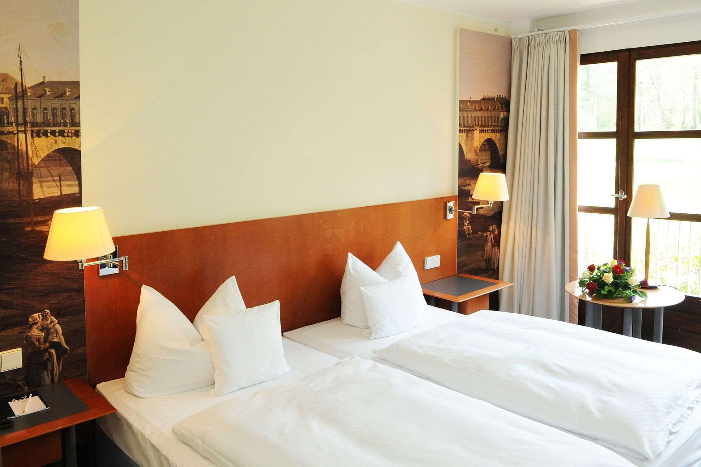 Een hotelkamer met twee bedden, een houten hoofdeinde, een nachtkastje met een lamp, een bijzettafeltje met een lamp, en een raam met gordijnen.