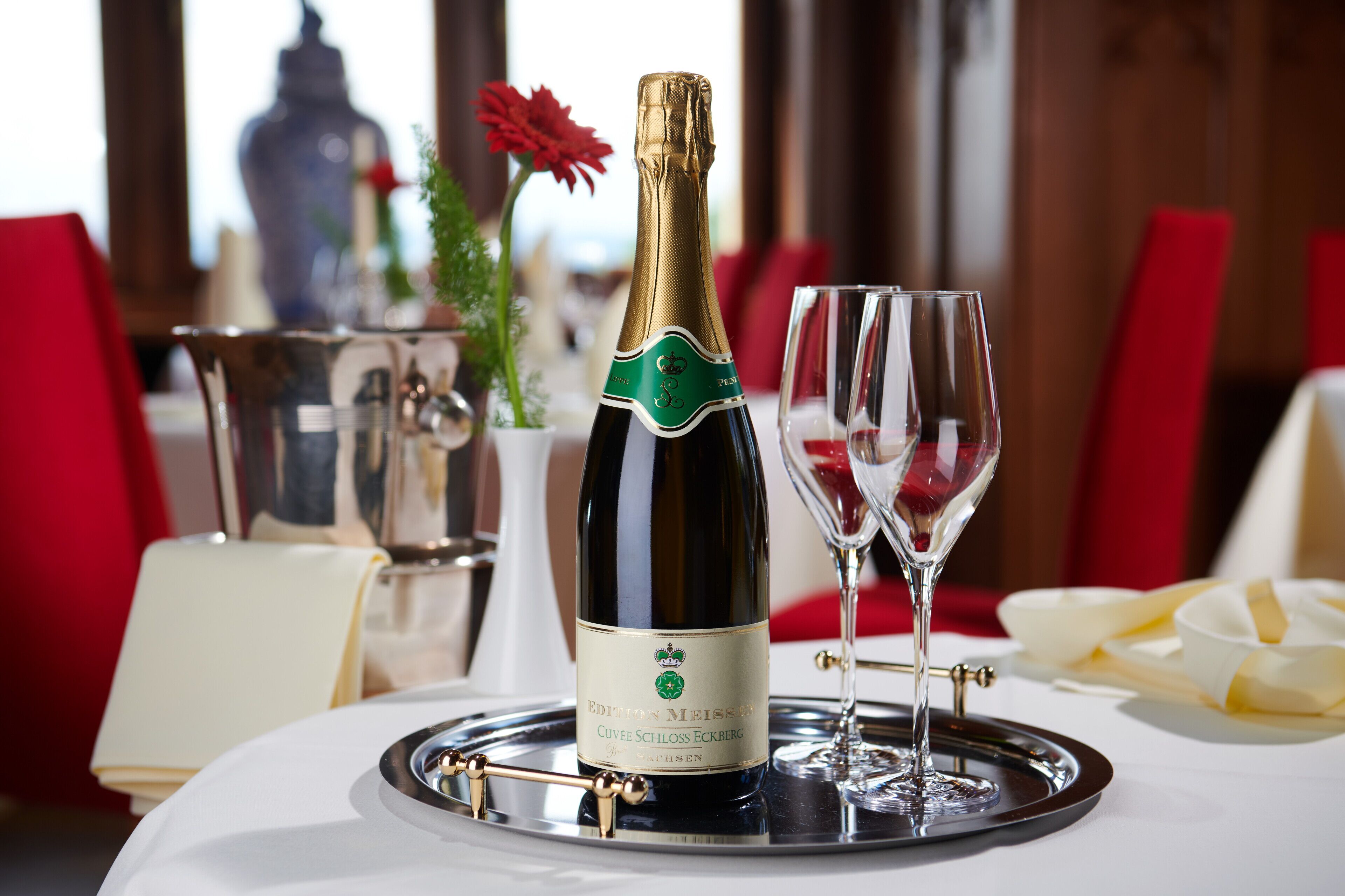 A bottle of Cuvée Schloss Eckberg wine on a tray with two glasses, set on a table with a white tablecloth and red chairs.