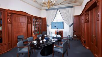 Uma sala de reuniões formal, com uma grande mesa de conferências, cadeiras, estantes de livros e um candelabro.