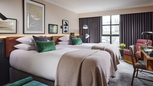 A hotel room with two beds, a desk, and a chair. There are framed artworks on the wall and a window with curtains. - The Alex (Dublin)