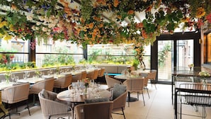 An indoor dining area with wicker chairs and tables, a hanging floral arrangement, and a view of greenery outside. - The Alex (Dublin)