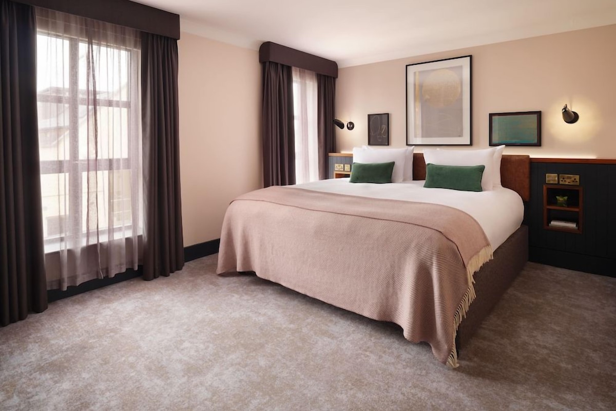 Une chambre d’hôtel avec un grand lit, deux tables de chevet, une commode, une fenêtre avec des rideaux et une lampe fixée au mur.