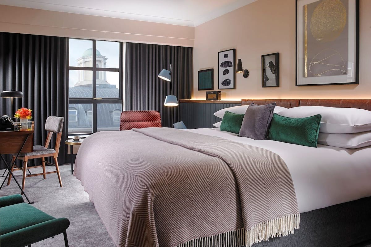 Habitación de hotel con una cama grande, un escritorio, una silla, una ventana con cortinas y obras de arte en las paredes.