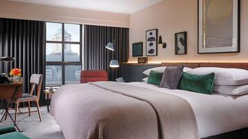 Habitación de hotel con una cama grande, un escritorio, una silla, una ventana con cortinas y obras de arte en las paredes.