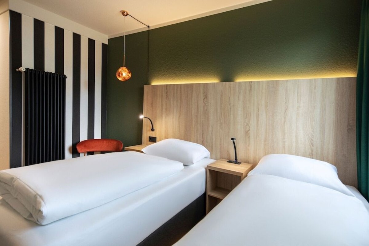 A hotel room with two beds, a red chair, a wooden headboard, and a hanging pendant light.