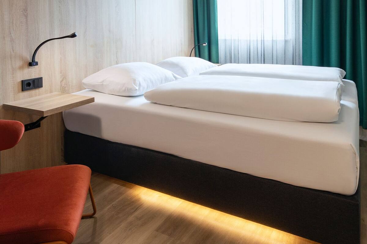 Ein ordentlich bezogenes Bett mit weißen Bettwäsche, ein Nachttisch aus Holz mit einer Lampe und ein roter Sessel in einem Hotelzimmer mit Holzboden und grünen Vorhängen.