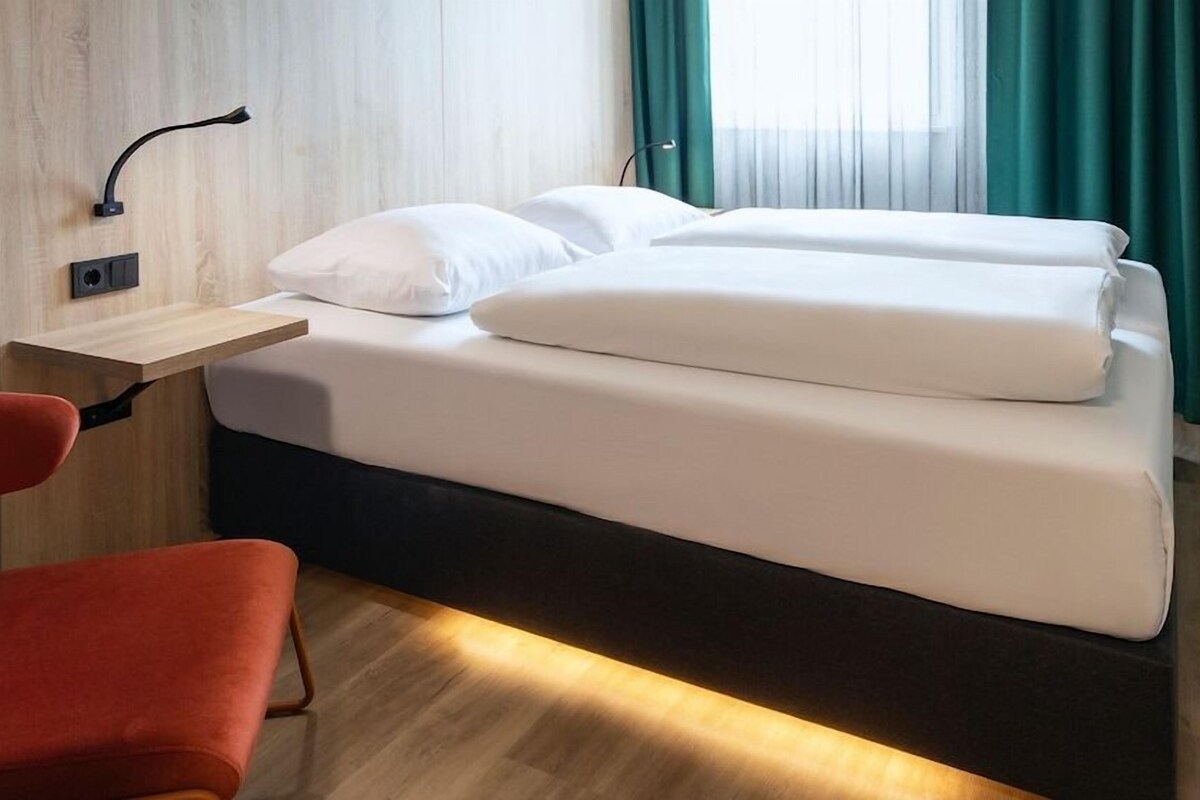 Una habitación de hotel con una cama, un escritorio, una silla y un ventanal con cortinas.