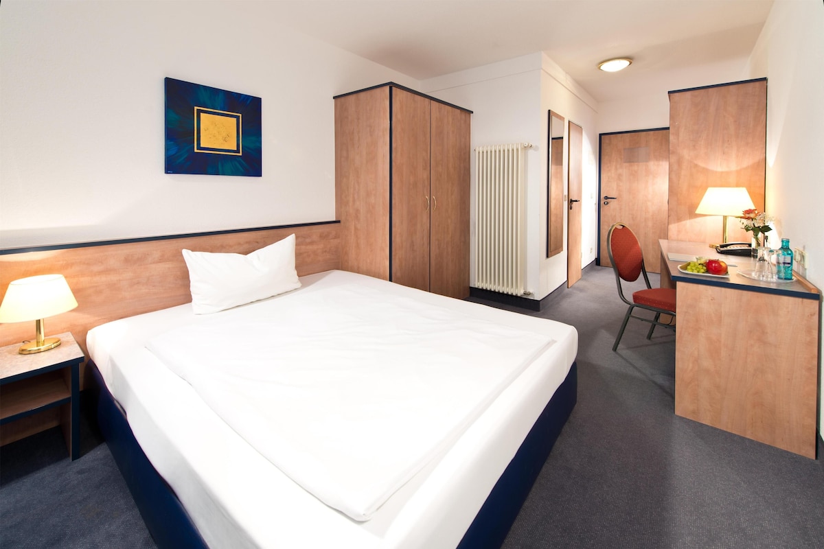 Quarto de hotel com uma cama grande, uma escrivaninha com cadeira, um radiador e um quadro emoldurado na parede.