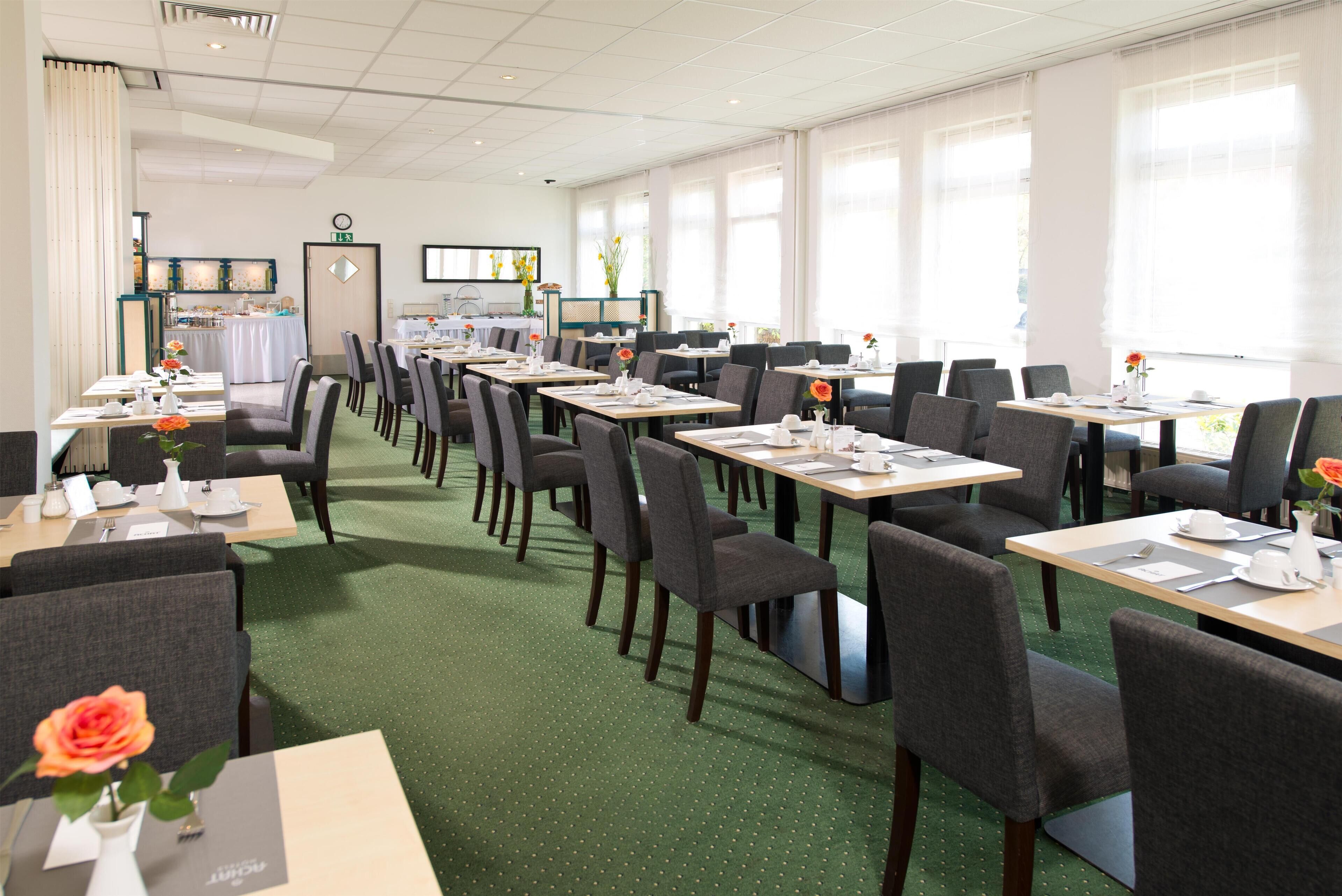 Ein Hotel-Speisesaal mit mehreren Tischen und Stühlen, einem grünen Teppich und großen Fenstern.