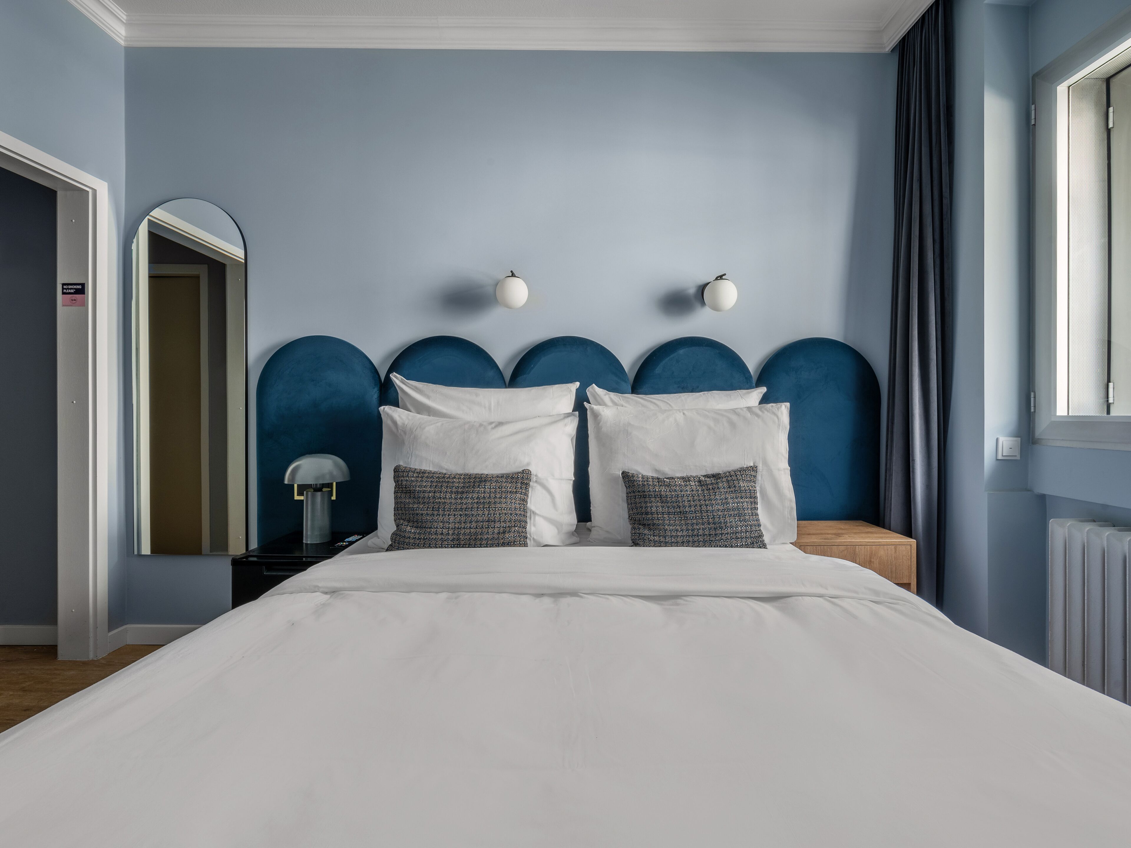 A bedroom with a bed, two bedside tables, a mirror, and a window with curtains.