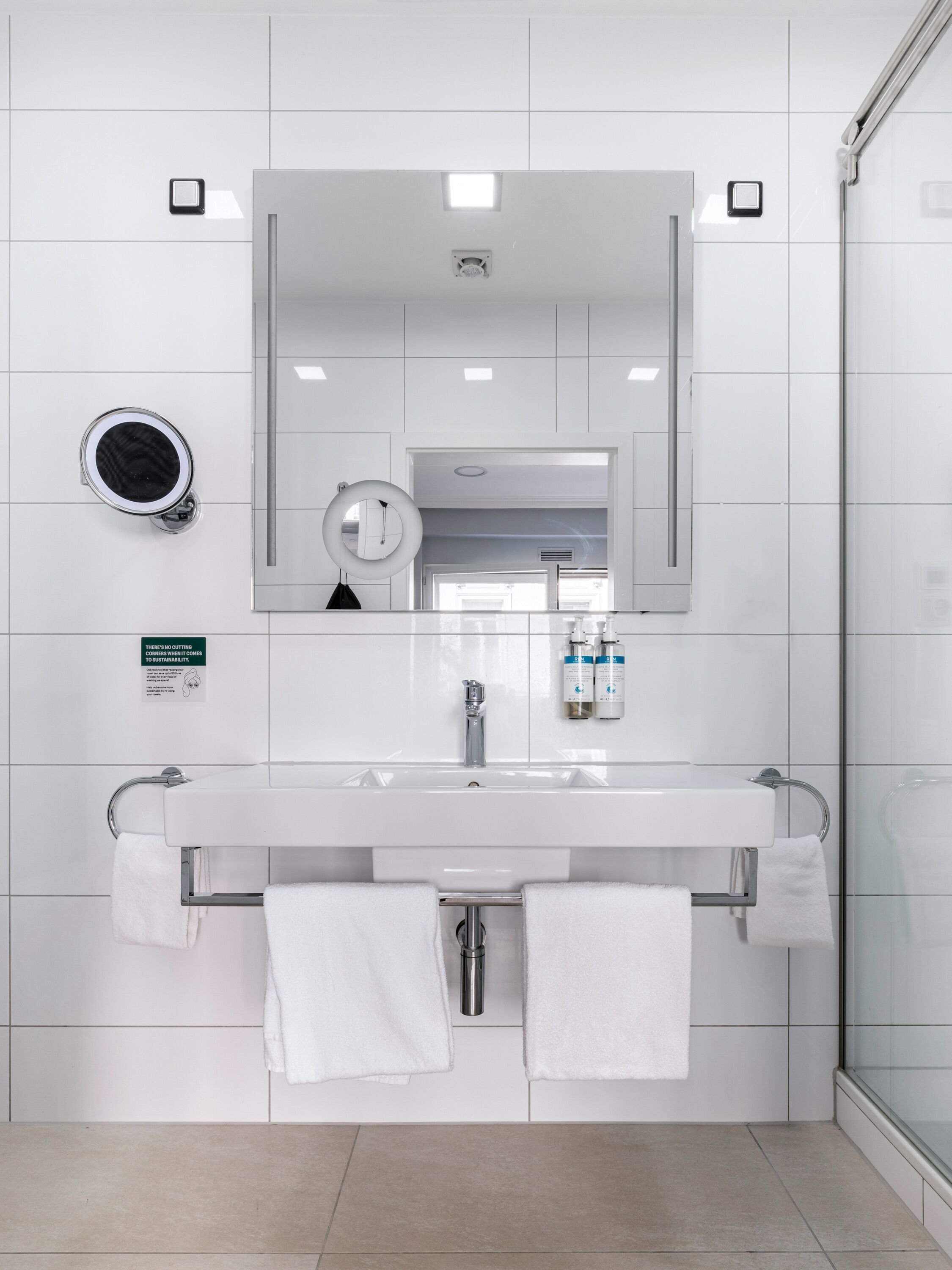 A modern bathroom with a white sink, mirror, and towel rack. There is a sign about sustainability on the wall.