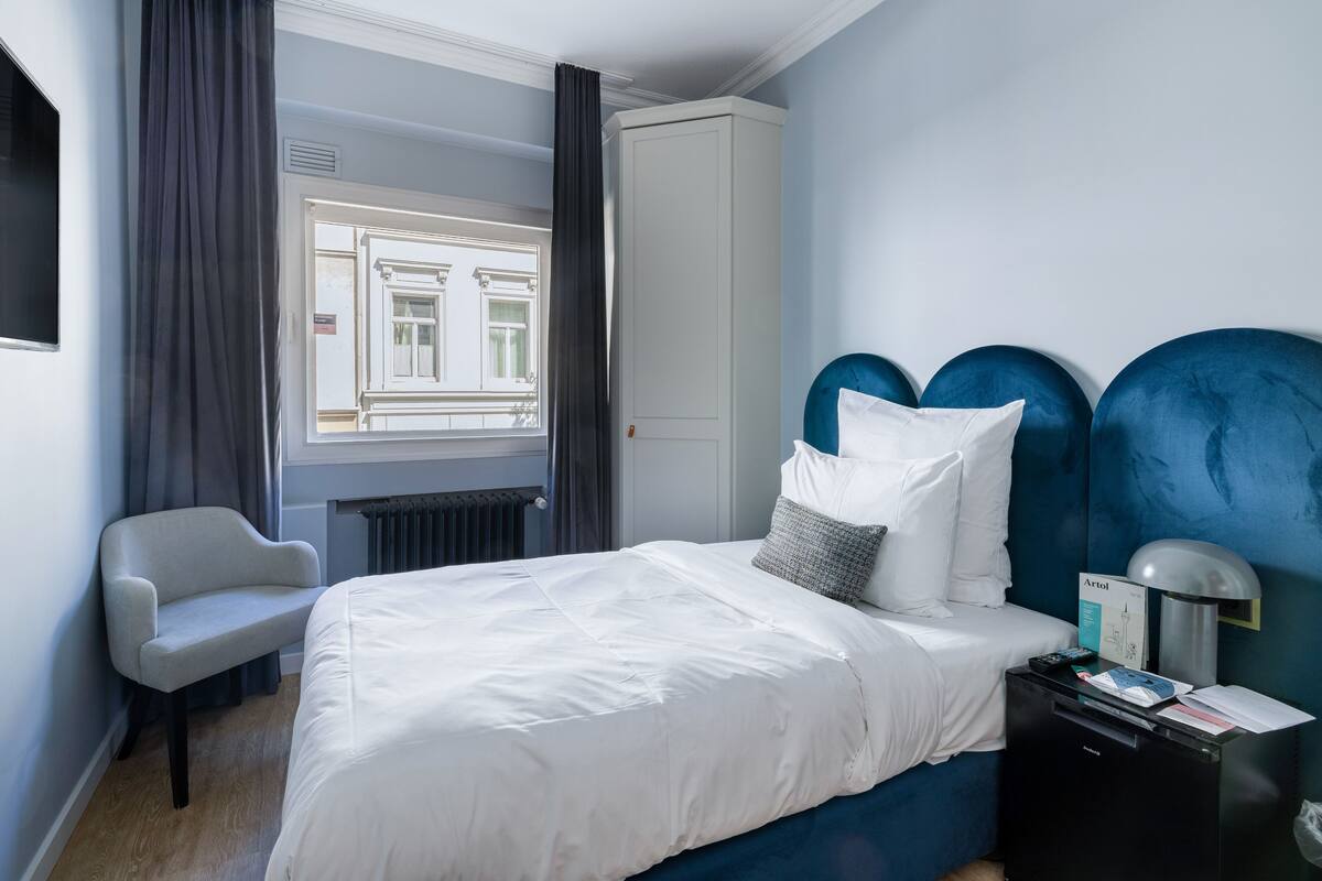 Une chambre d’hôtel avec un lit, une chaise, un bureau avec une lampe et une vue sur un immeuble par la fenêtre.