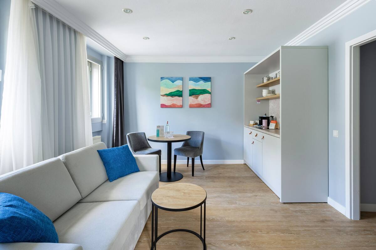 Un salon moderne comprenant un canapé, des fauteuils, une petite table ronde et un coin cuisine avec des étagères.