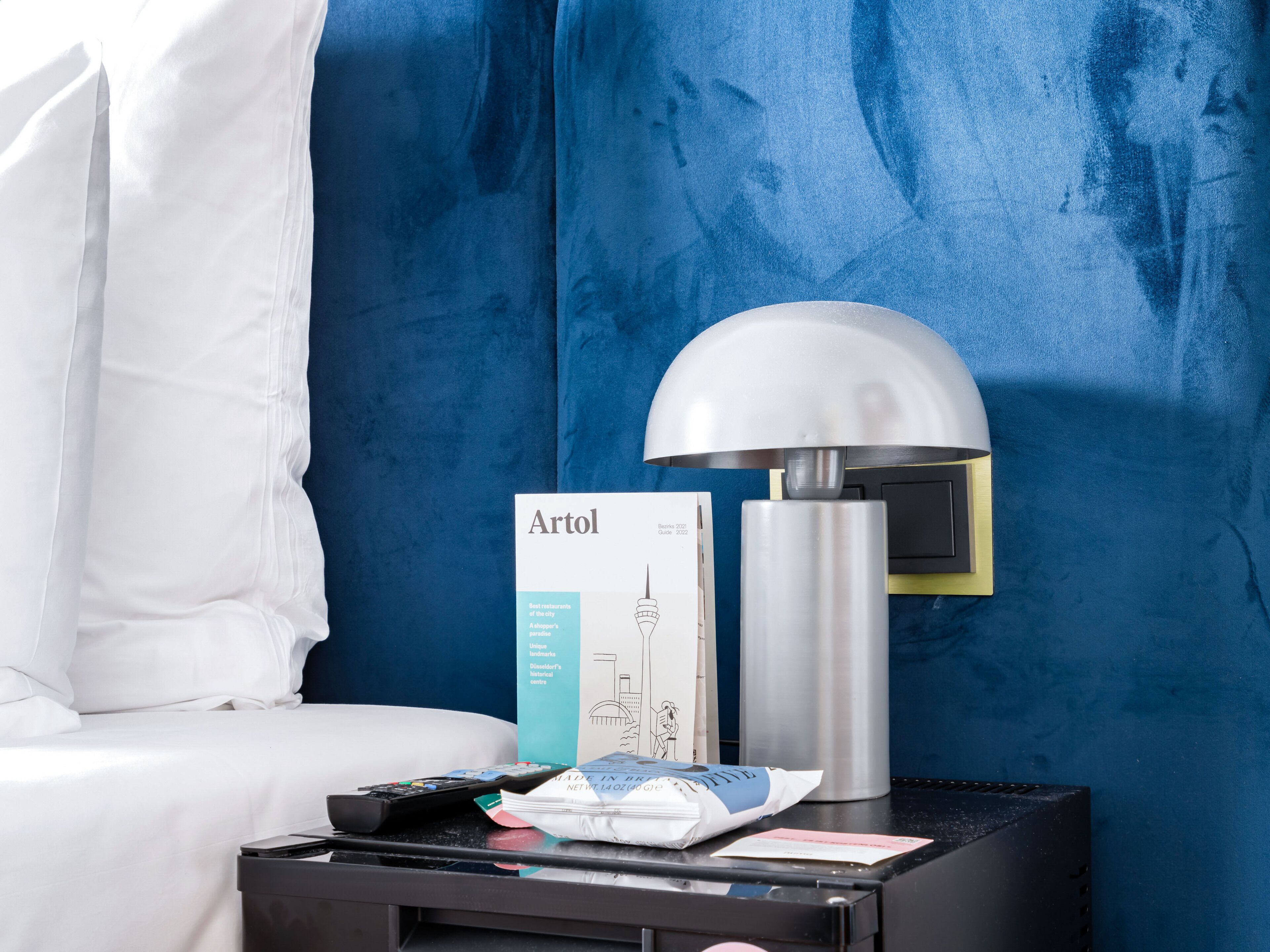 A hotel room with a bed, a nightstand, a lamp, a remote control, a brochure, and a bag.