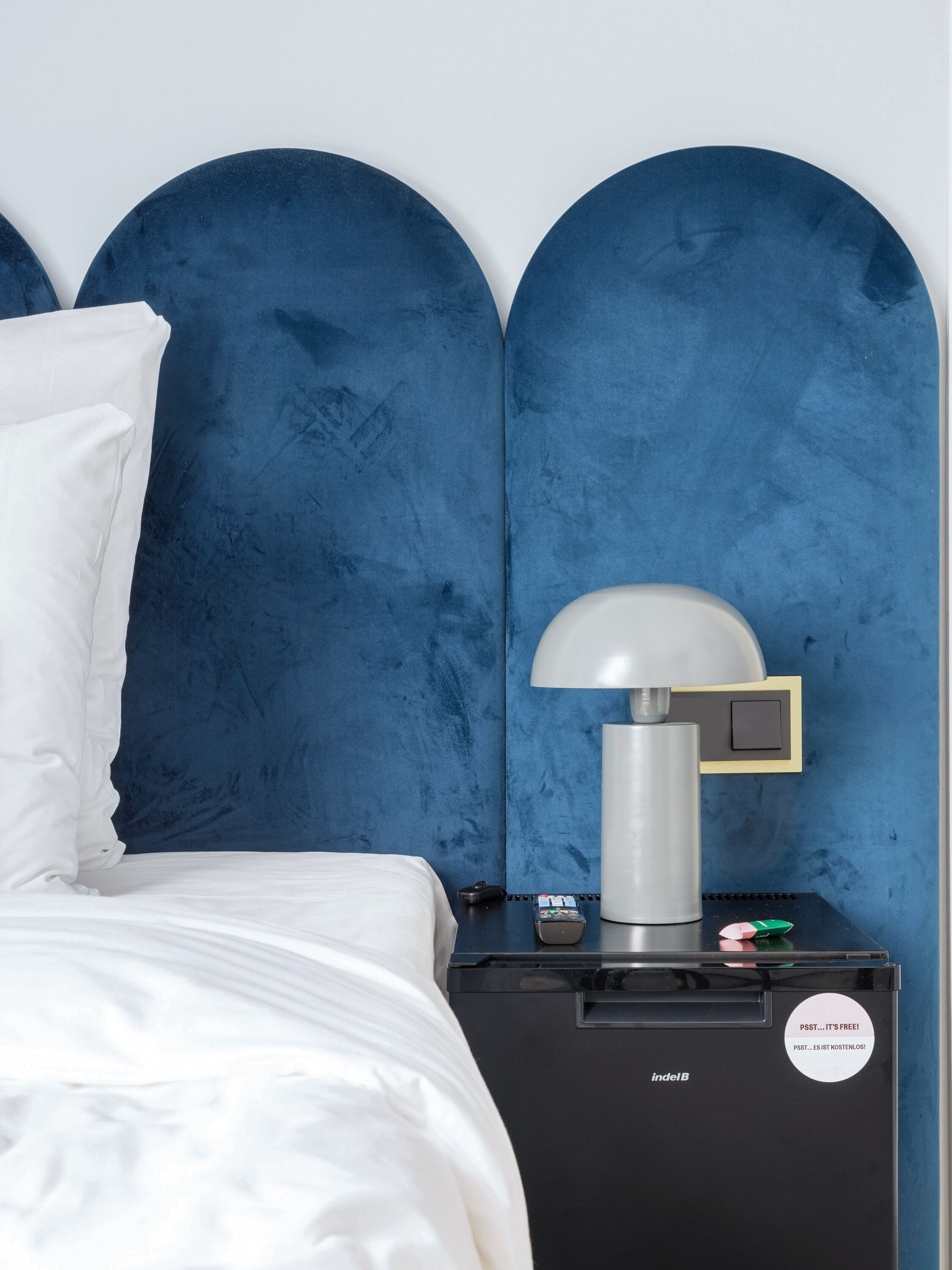A hotel room with a bed, a nightstand with a lamp, and a minibar.