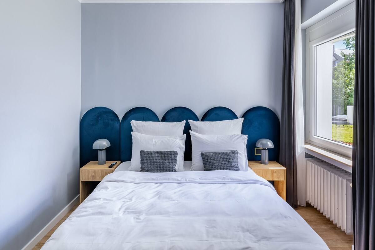 Une chambre à coucher avec un lit, deux tables de chevet, une fenêtre avec des rideaux et un radiateur.