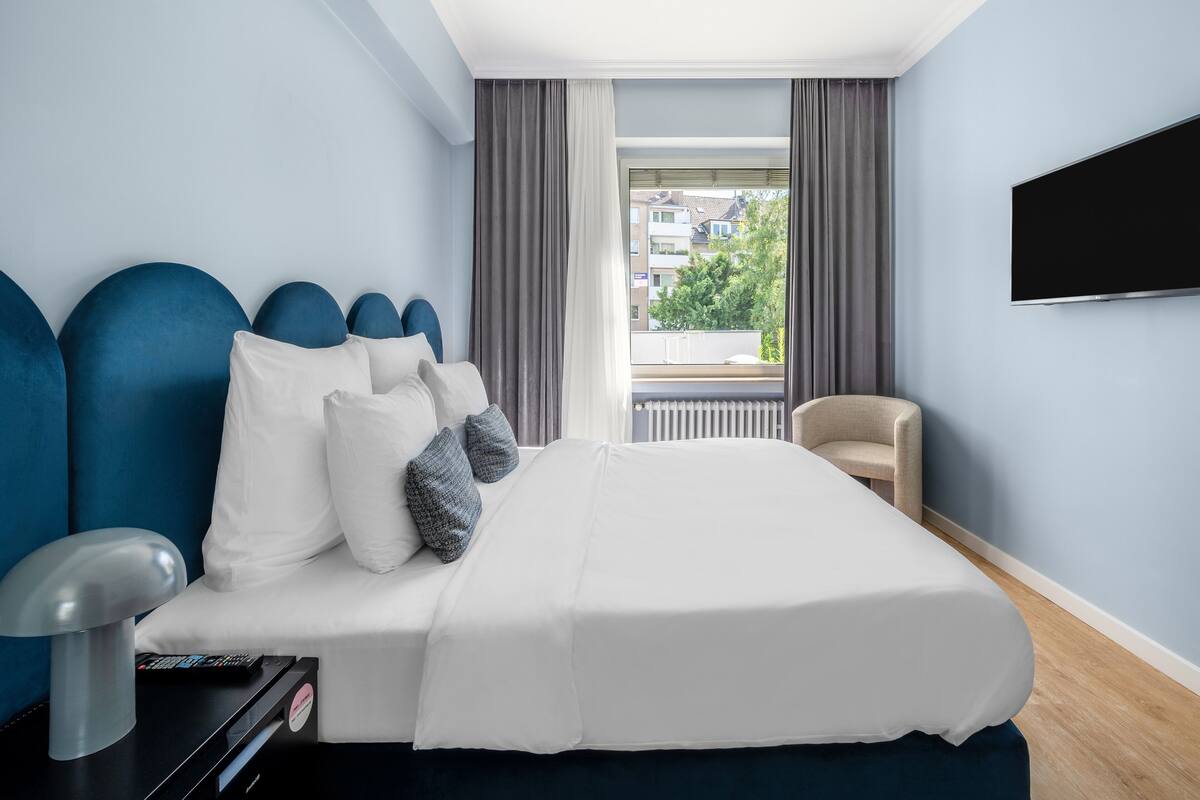 Une chambre d’hôtel moderne avec un grand lit, un téléviseur fixé au mur et une vue sur l’extérieur par une fenêtre.