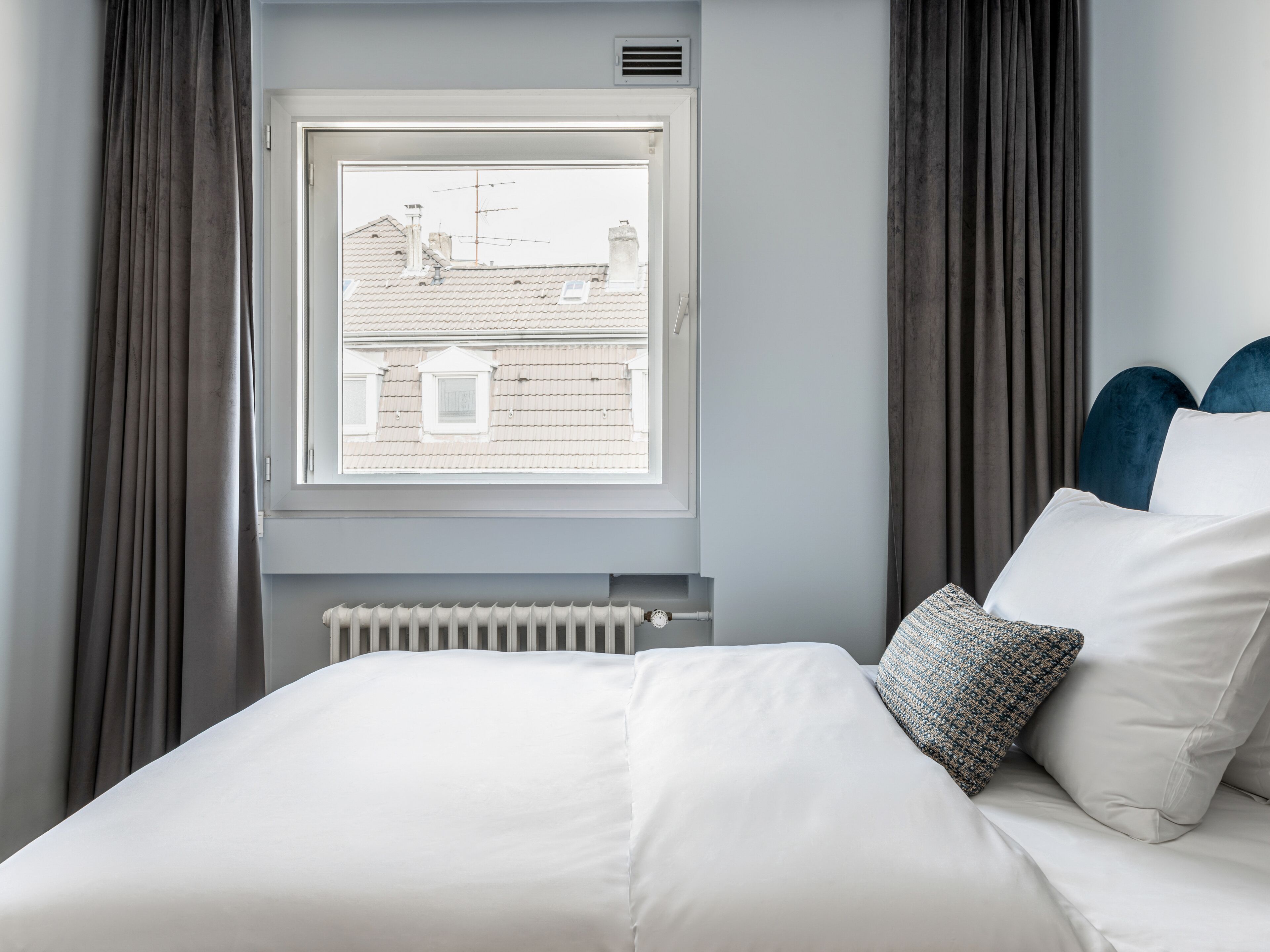 Un dormitorio con una cama, una ventana con vistas a edificios y cortinas grises.