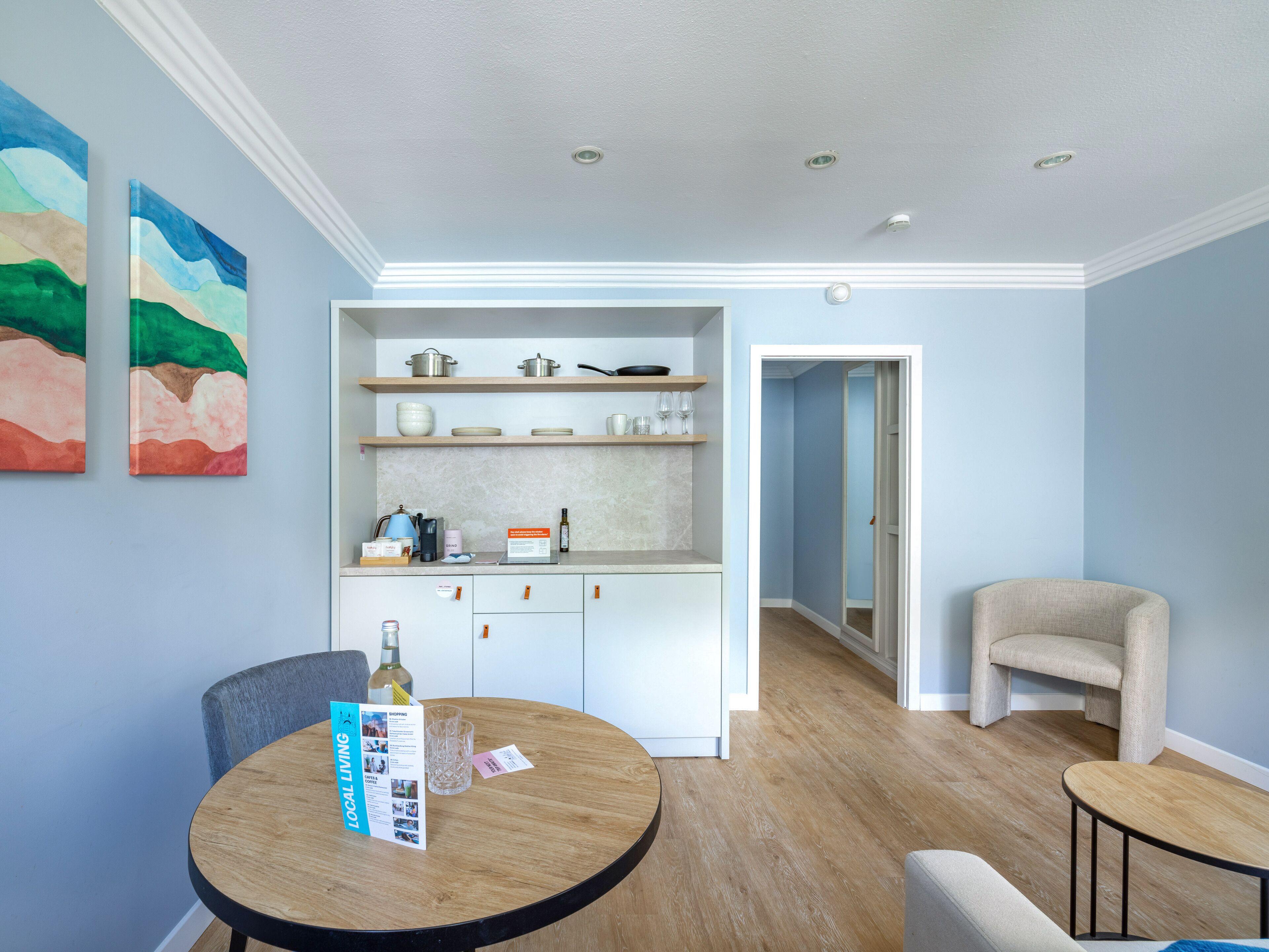 A modern kitchen with white cabinets, a round wooden table, and a chair. There are two abstract paintings on the wall.