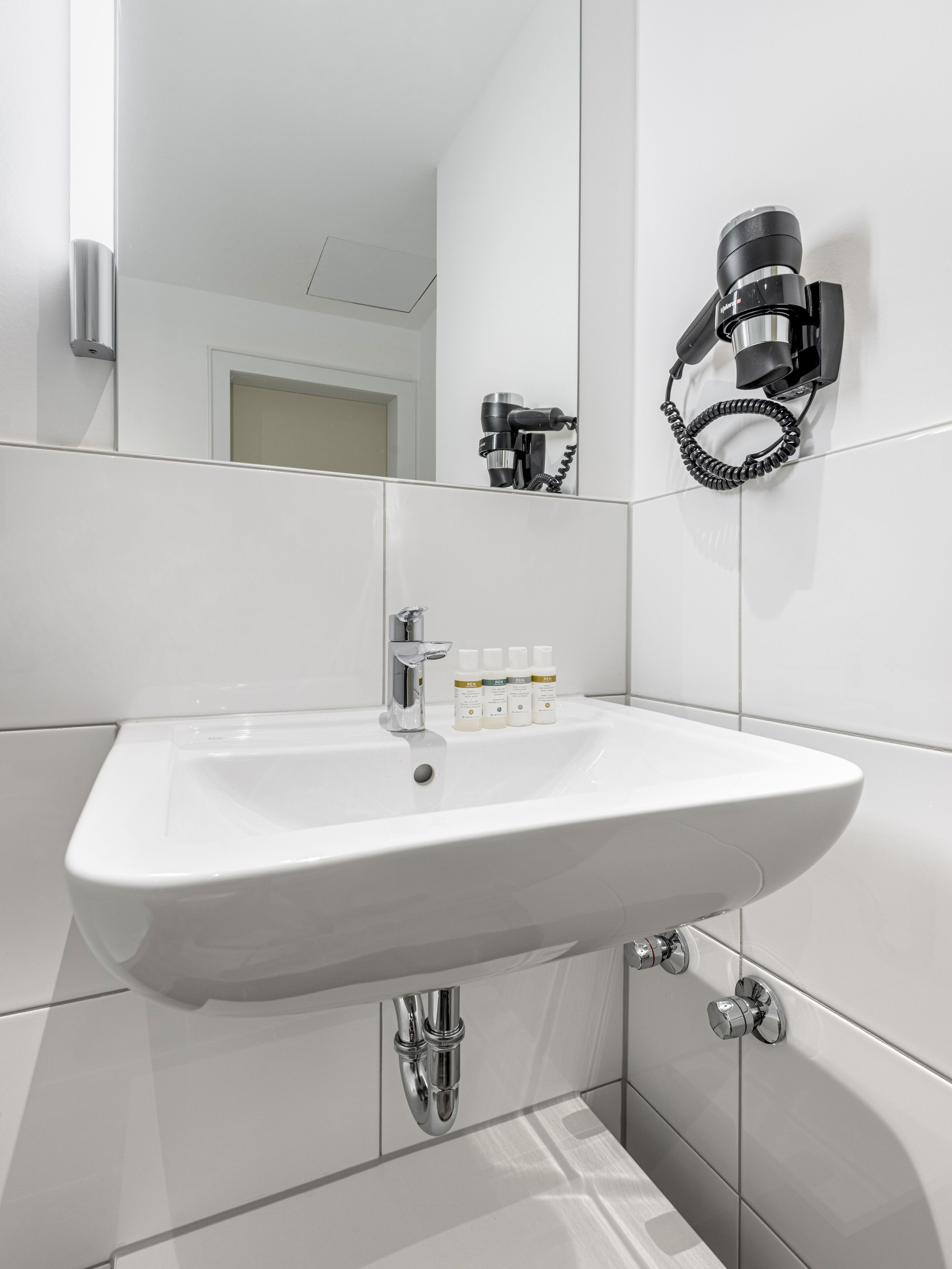 Un baño moderno con un lavabo blanco, un secador de pelo de mano montado en la pared y un espejo.