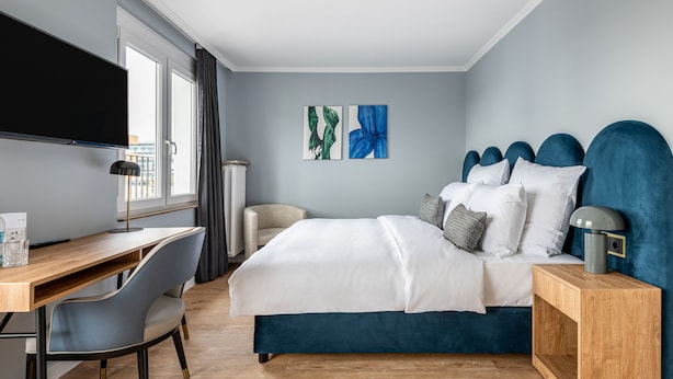 Ett modernt hotellrum med en säng, ett skrivbord och en stol. Det finns en TV som är monterad på väggen och konstverk på väggen.