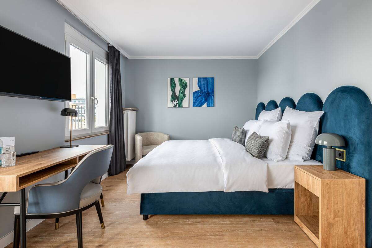 Une chambre d’hôtel moderne, équipée d’un lit, d’un bureau et d’une chaise. Un téléviseur est fixé au mur, et des œuvres d’art décorent également ce même mur.