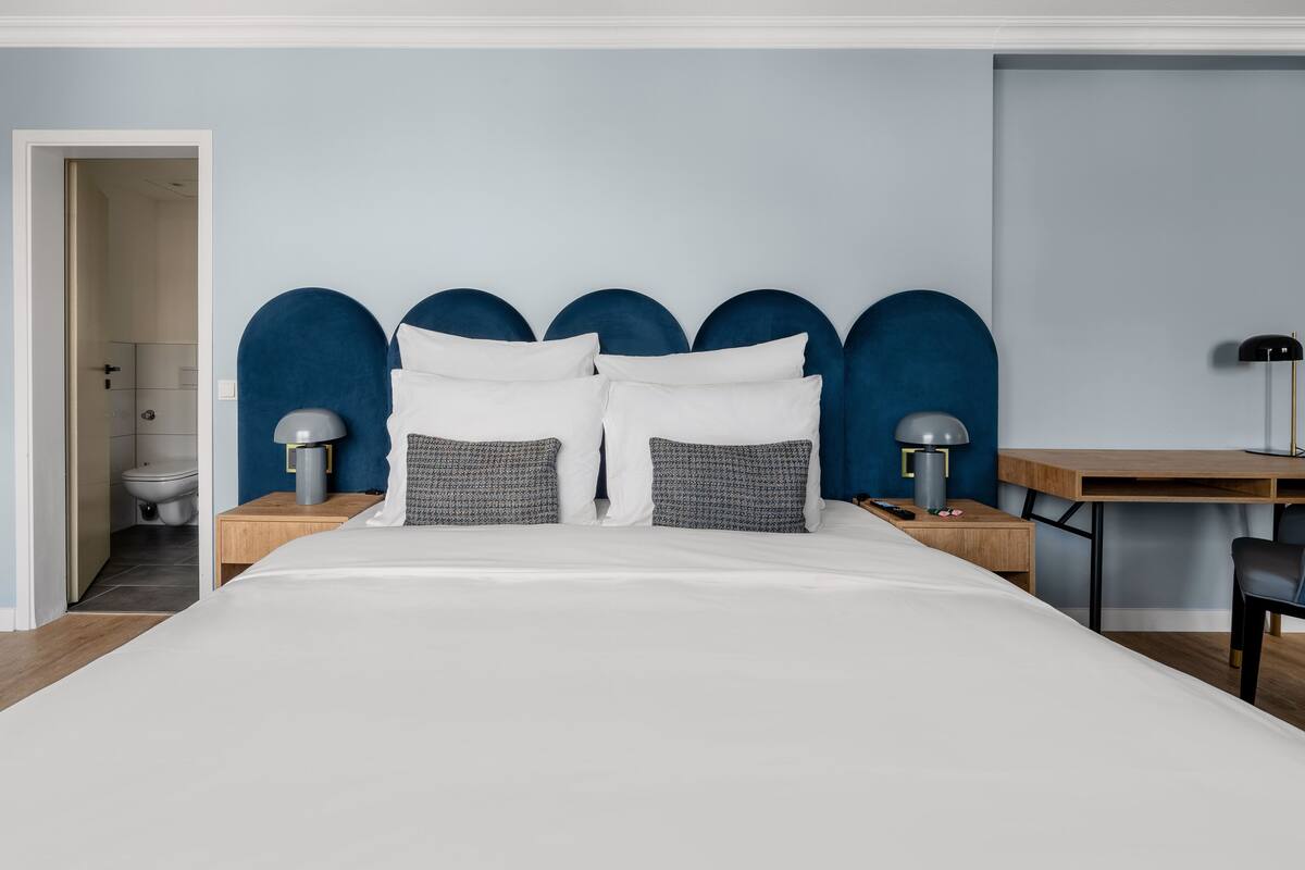 Une chambre à coucher moderne avec un grand lit, un bureau et une chaise. On aperçoit une salle de bain au fond.