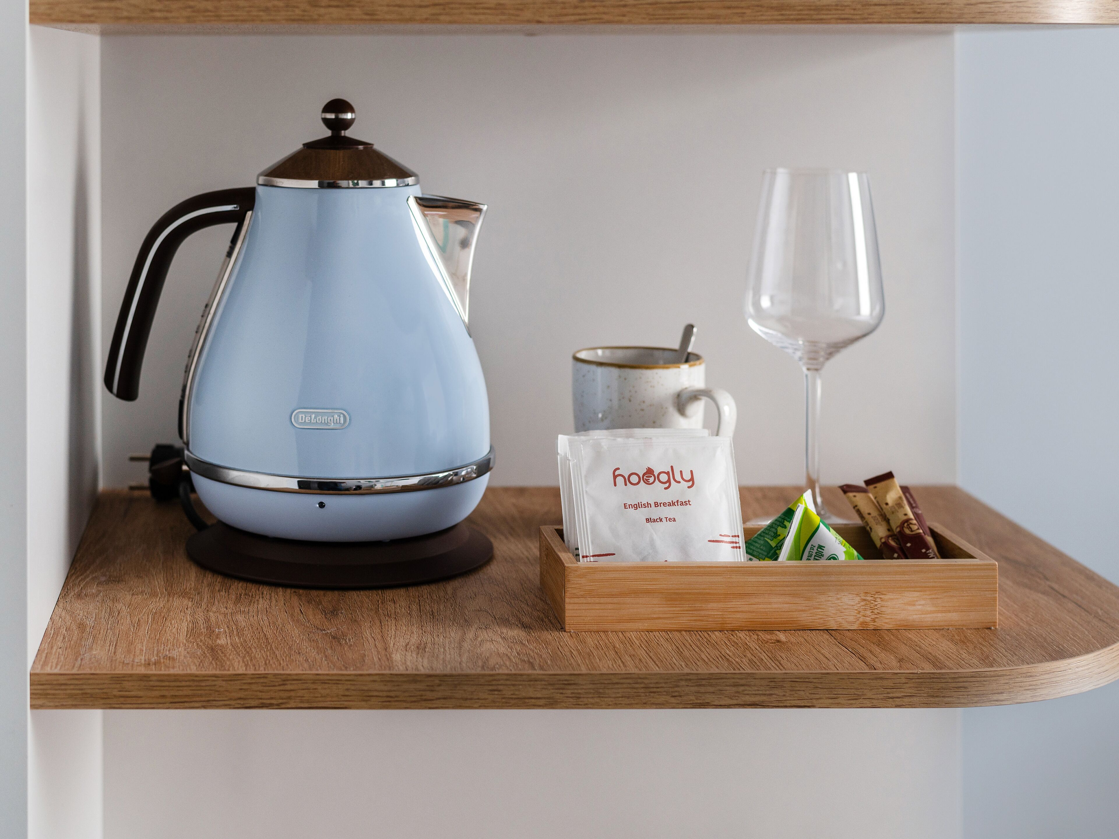 Une bouilloire électrique bleue, une tasse en verre, un sachet de thé noir Hoagly et un verre d’eau, le tout posé sur une étagère en bois.