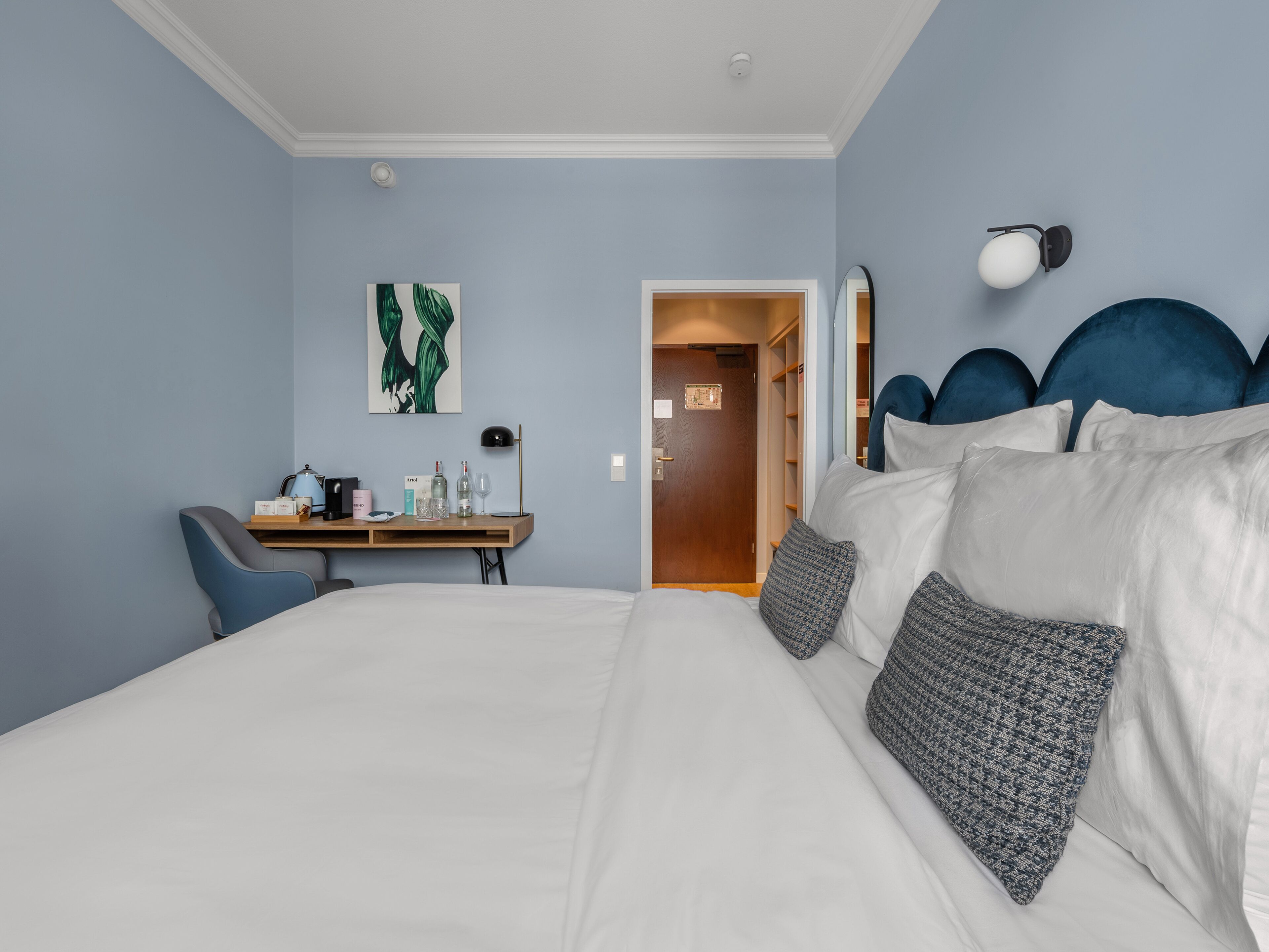 A hotel room with a large bed, a desk with a chair, a wardrobe, and a mirror on the wall.