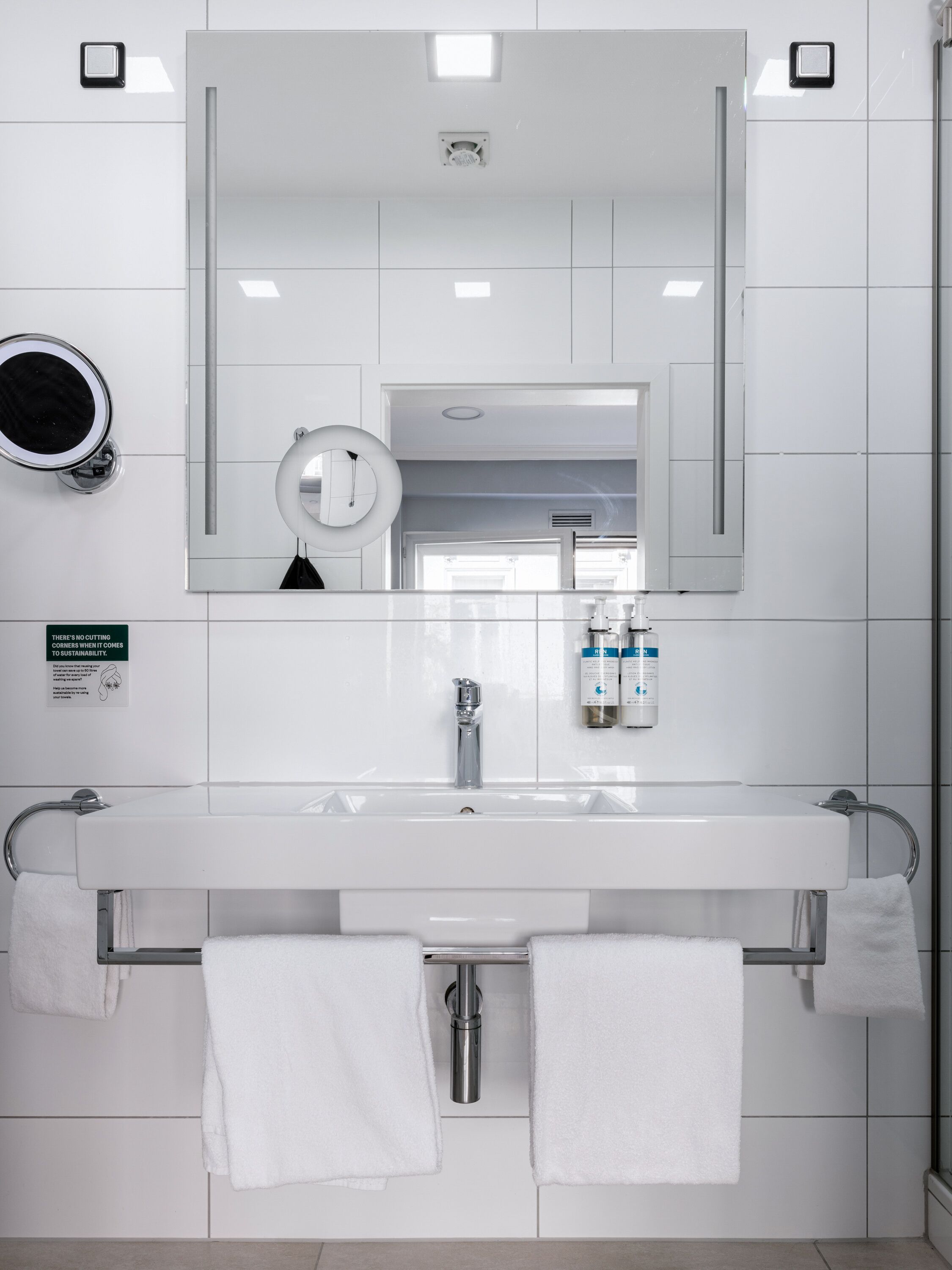 A modern bathroom with a white sink, two towel racks, a mirror, and a sign about sustainability.