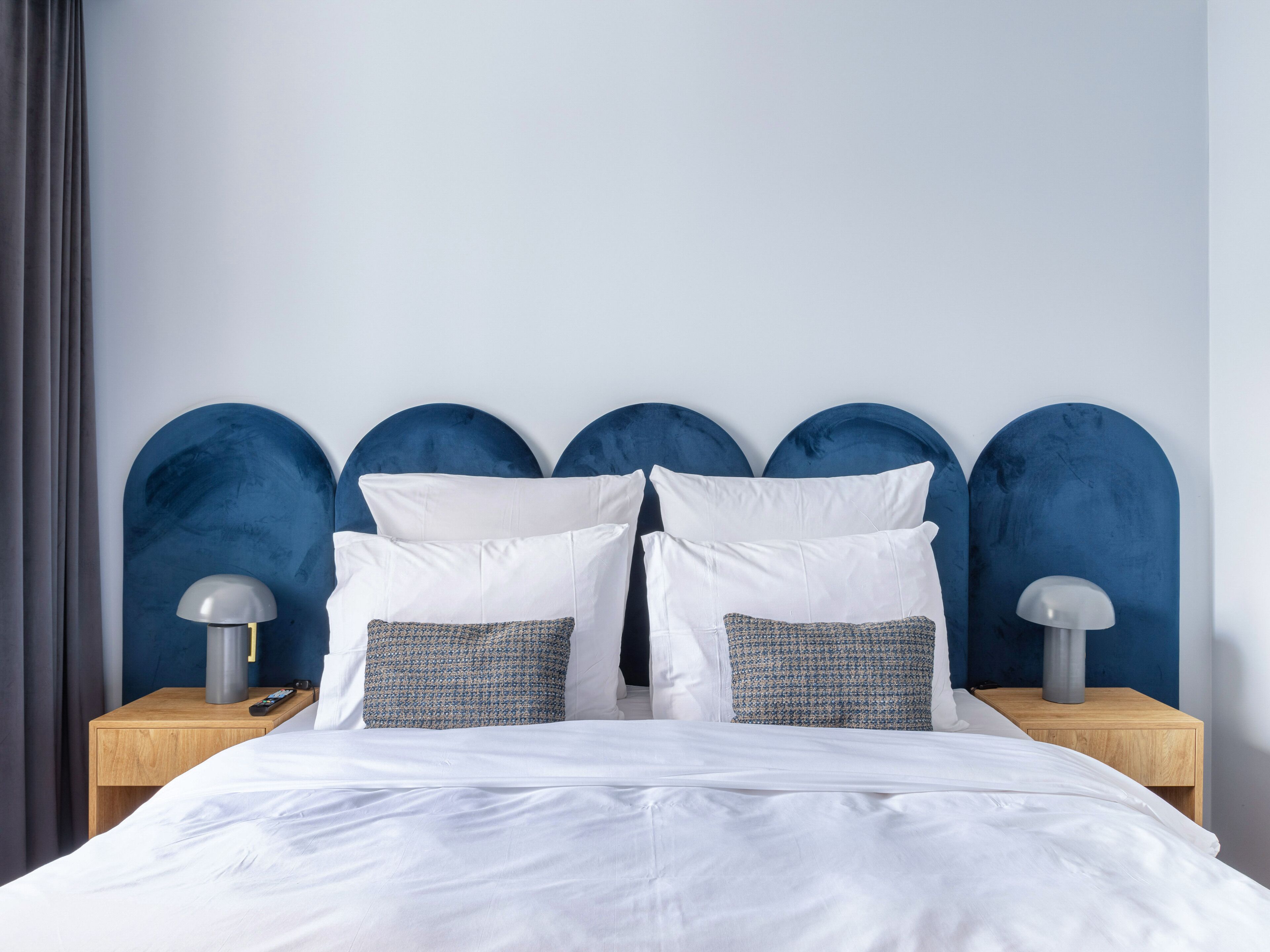 A neatly made bed with white linens, two white pillows, and two patterned pillows. There are two bedside tables with lamps and a remote control on one of them. The headboard is blue with a unique curved design.