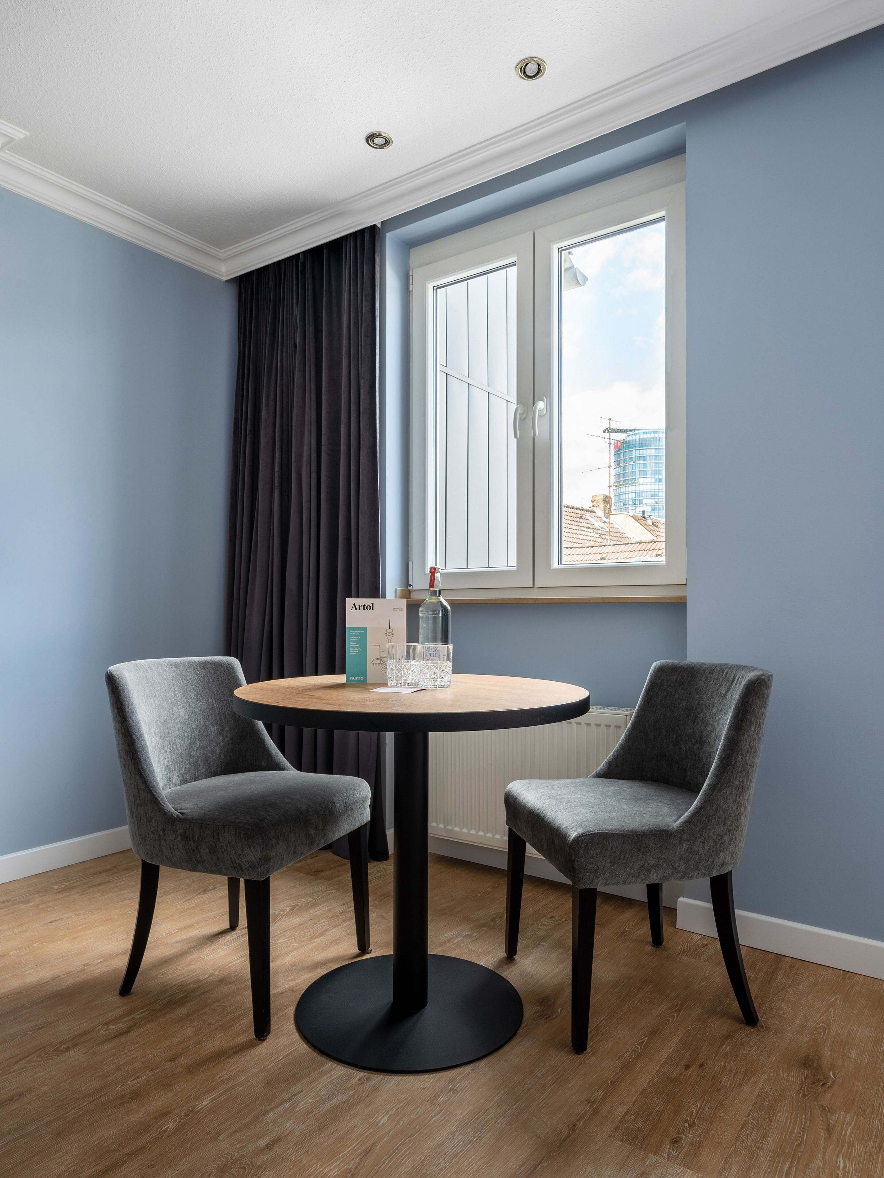 A room with a round table, two grey chairs, a window with curtains, and a radiator.