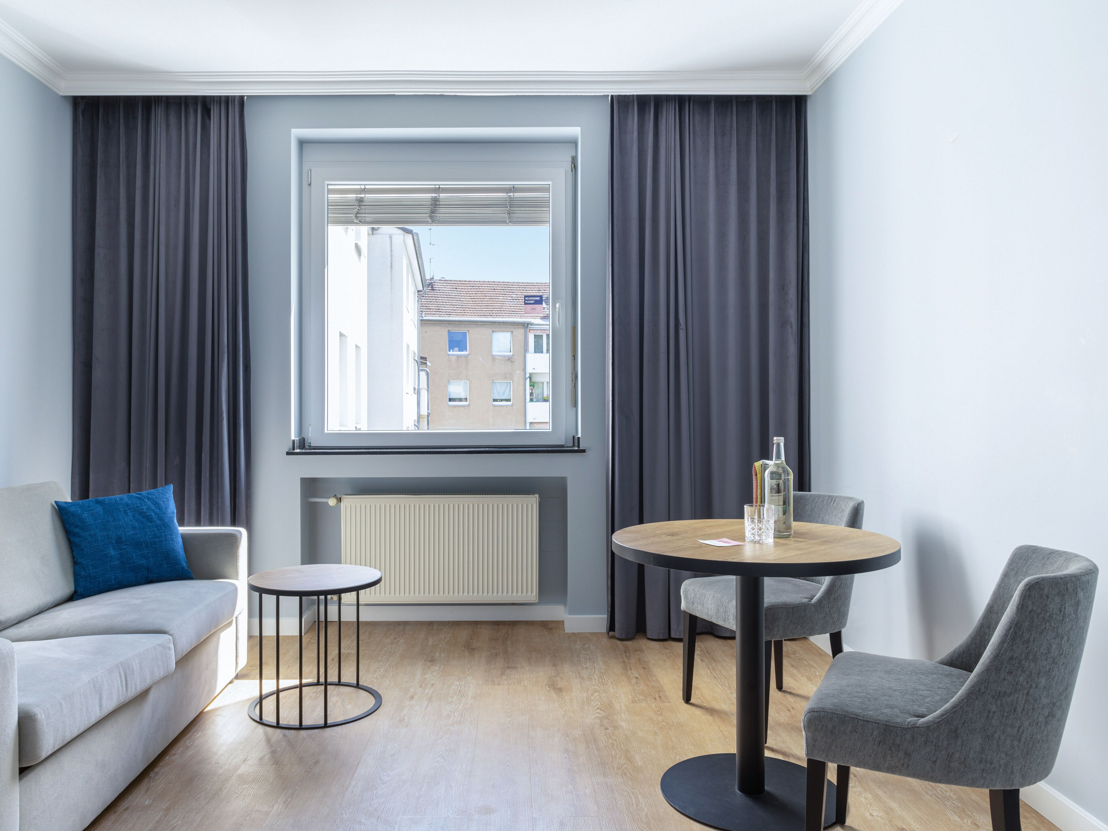 A modern room with a grey sofa, a round table with two chairs, a window with blinds, and a radiator.