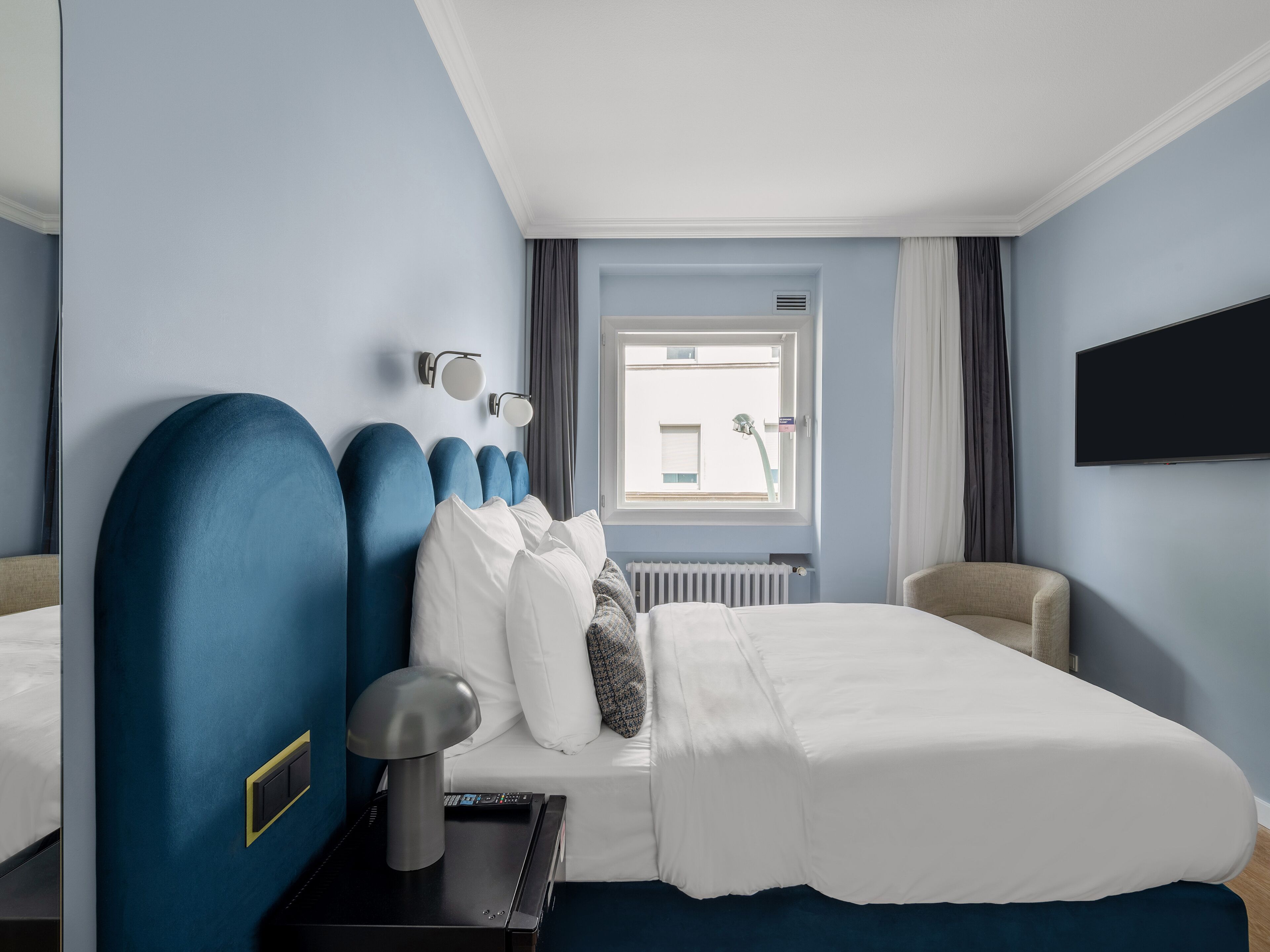 A hotel room with a bed, a chair, a desk with a lamp, a TV, and a window with curtains.
