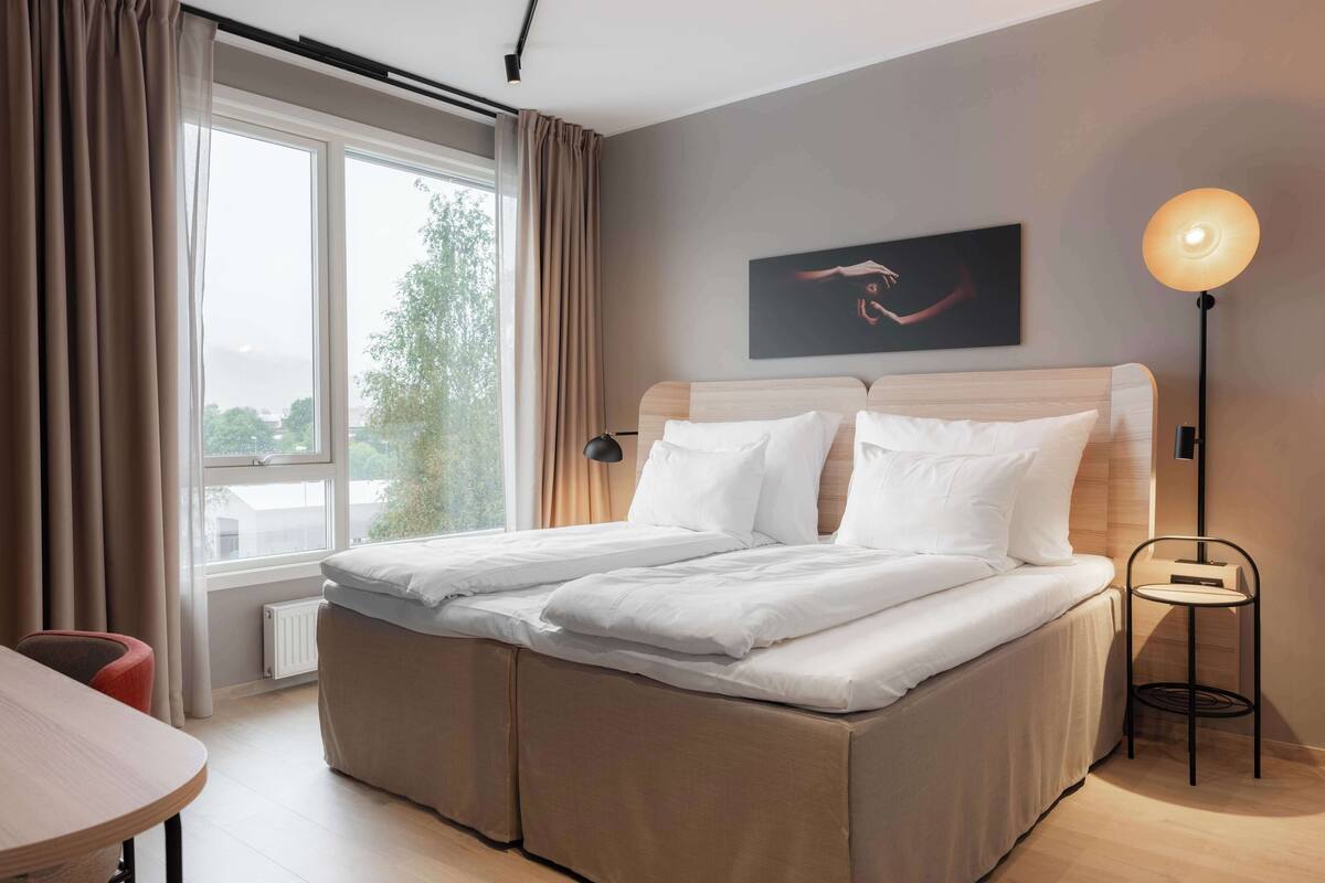 Ein modernes Hotelzimmer mit einem großen Bett, einem Nachttisch, einer Lampe und einem Fenster mit Vorhängen.