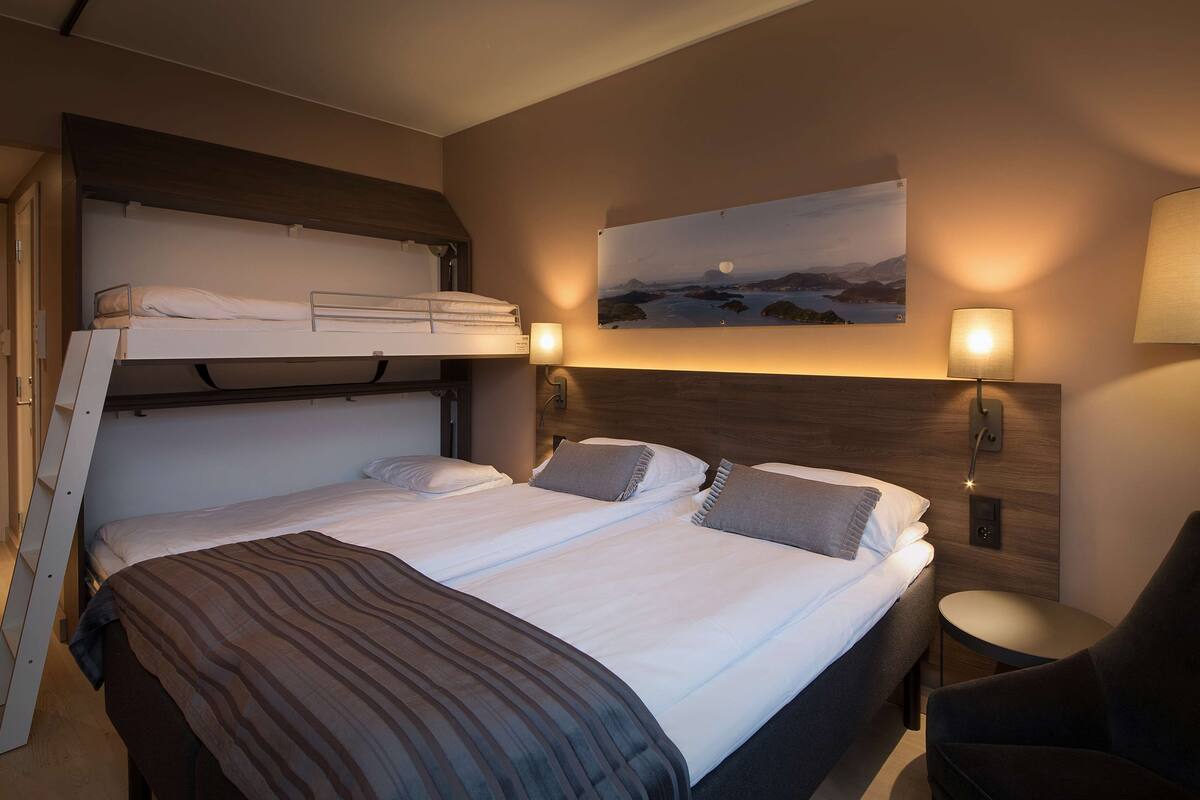 Ein Hotelzimmer mit Etagenbett, einem Einzelbett, einem kleinen runden Tisch, einem Stuhl und einer Wandlampe.