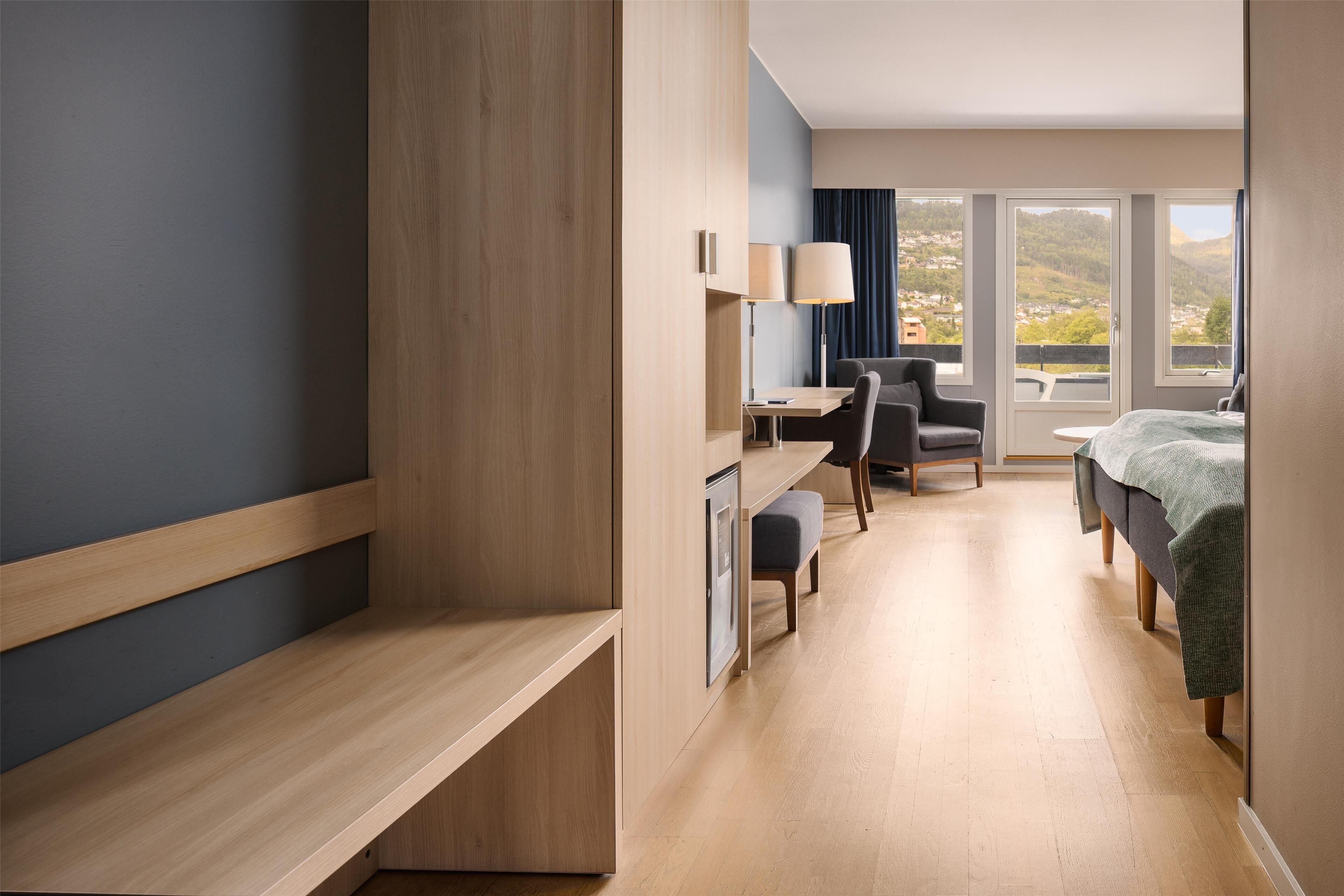 Una habitación de hotel moderna con un escritorio de madera, una cama, una silla y vistas a la ciudad a través de amplias ventanas.