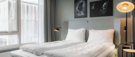 A modern hotel room with a large window, two beds, a nightstand, and a lamp.