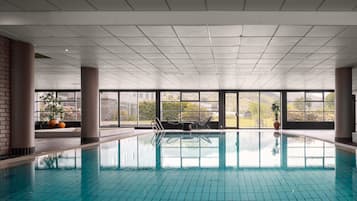 Une piscine intérieure à l’eau claire, entourée de grandes fenêtres qui offrent une vue sur la verdure et les bâtiments.