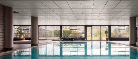 An indoor swimming pool with clear water, surrounded by large windows offering views of greenery and buildings.